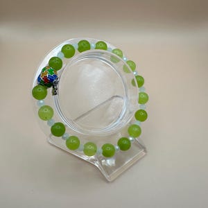 May include: A bracelet with large, round, lime green beads and smaller, clear beads. A decorative charm with colourful enamel accents hangs from the bracelet. The bracelet is displayed on a clear, angled stand.