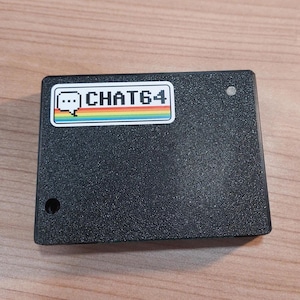 May include: A black rectangular device with a textured surface. A sticker with the text "CHAT64" in white, a speech bubble icon, and a rainbow stripe is affixed to the top. There are two small holes on the device.