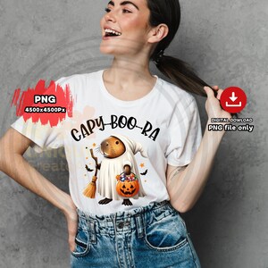 以下が含まれることがあります： 「Capy-Boo-Ra」という文字がプリントされた白いTシャツ。幽霊に扮したカピバラの漫画が、お菓子の入ったカボチャのバケツとほうきを持っています。