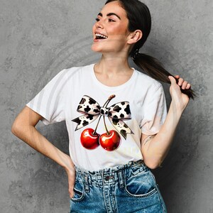 Op de afbeelding: Wit T-shirt met een zwart-witte koe-print strik en twee rode kersen.