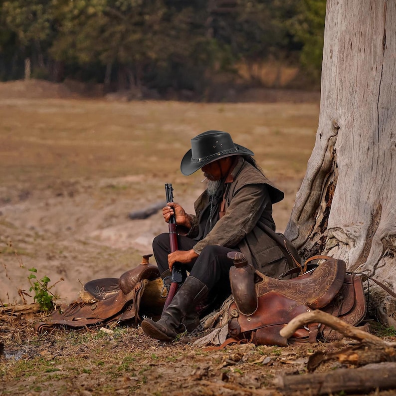 May include: A man wearing a black cowboy hat sits next to a tree with a rifle in his hand. He is wearing a brown jacket and jeans. There are two saddles in the background.
