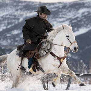May include: A cowboy wearing a brown coat and a black hat rides a white horse through a snowy landscape.