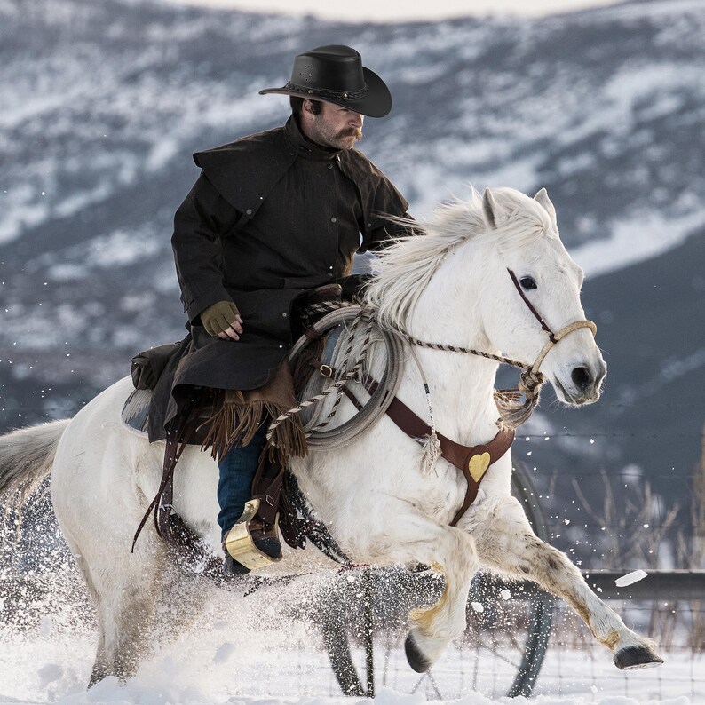 May include: A cowboy wearing a brown coat and a black hat rides a white horse through a snowy landscape.