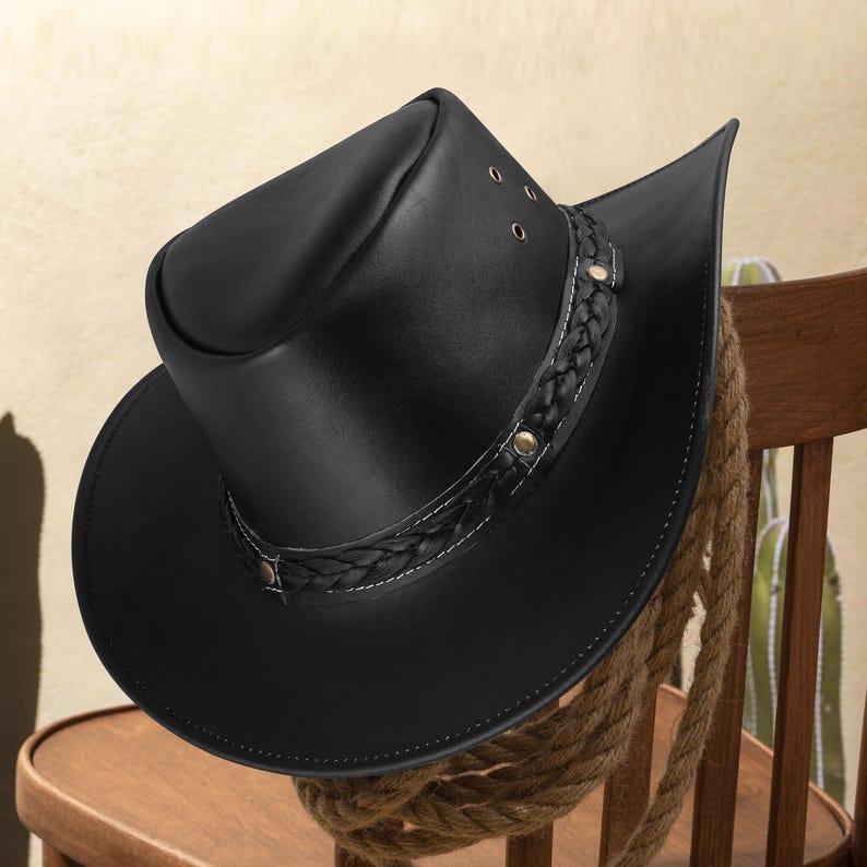 May include: A black leather cowboy hat with a braided band and a small brim. The hat is sitting on a wooden chair.