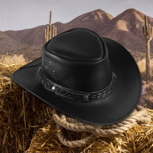 May include: A black leather cowboy hat with a braided band and a wide brim. The hat is sitting on a bale of hay.