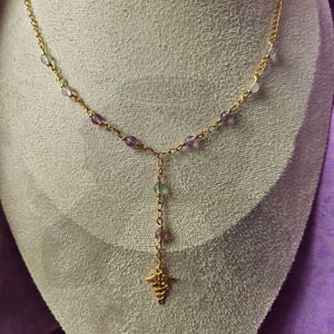 Può includere: Una collana color oro con una delicata catena, caratterizzata da piccole perline colorate nelle tonalità del viola e del verde. Un ciondolo con simbolo medico dorato pende dal centro, creando un design unico ed elegante.
