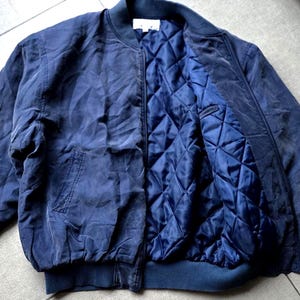 May include: A navy blue bomber jacket featuring a ribbed collar, cuffs, and hem. The jacket has a front zipper closure and a quilted interior lining in a matching navy blue tone. A white label is visible at the inner neckline of the garment.