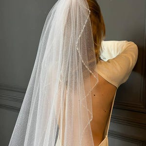 May include: A white bridal veil with a delicate pattern of small dots and a beaded trim. The veil is made of soft tulle and is designed to be worn over the head.