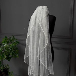 May include: A white, two-tiered bridal veil with a beaded edge. The veil is made of a sheer, shimmering fabric and is displayed on a black mannequin. The background is a dark gray wall with a decorative trim and a potted plant.