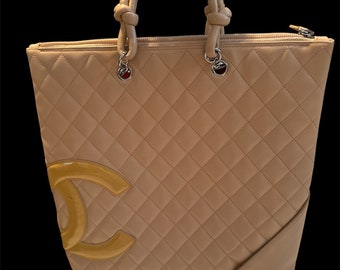 May include: A beige quilted leather tote bag with a large gold Chanel logo. The bag has two top handles and a zipper closure.