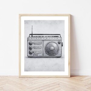 May include: A framed grayscale print of a vintage radio. The radio has a dial, antenna, and buttons labeled "GOOD NEWS", "BAD NEWS", and "FAKE NEWS". The artwork is in a minimalist style, with a light wooden frame.