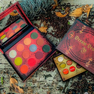 Può includere: Una palette di ombretti rossa e dorata con le parole "Dream Demon" sulla copertina. La palette contiene 12 tonalità, tra cui rosso, arancione, verde e marrone. La palette è aperta e le tonalità sono visibili. La palette è su una superficie di legno con foglie sparse intorno.