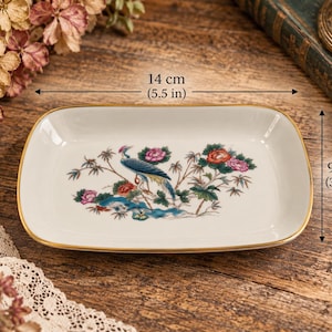 Peut inclure: Un plateau rectangulaire en porcelaine blanche avec un bord doré, orné d'une illustration centrale représentant un oiseau bleu parmi des fleurs colorées et un feuillage vert. Le plateau mesure 14 cm de large et 9 cm de haut.