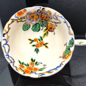 Puede incluir: Una taza de té de cerámica blanca con un diseño floral. La taza tiene un borde festoneado y un asa. El diseño floral presenta flores naranjas, amarillas, verdes y azules.