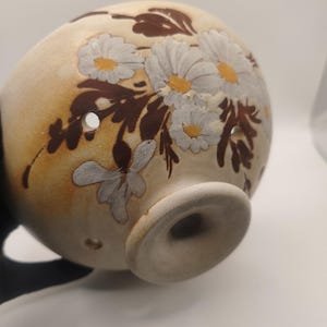 May include: A spherical ceramic vessel with a beige base, featuring hand-painted white daisy flowers with yellow centers and dark brown foliage. The object has a circular pedestal base and several small, evenly spaced circular holes along its side.