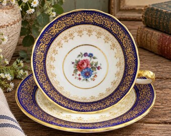 May include: A porcelain teacup and saucer set featuring a deep cobalt blue border with intricate gold filigree and floral patterns. The center of the cup displays a small, colorful bouquet of pink, blue, and green flowers on a white background.