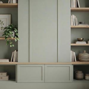 Puede incluir: Una unidad de pared integrada de color verde salvia con estantes y armarios de madera clara. Los estantes están llenos de varios libros, una lámina botánica enmarcada, jarrones de cerámica, recipientes con tapa y plantas verdes en macetas.