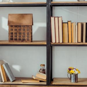 Könnte beinhalten: Ein schwarzes Metallregal mit Holzablagen, das verschiedene Bücher, ein braunes, hausförmiges Dekorationsobjekt, ein kleines Glasgefäß und einen silbernen Metallbehälter mit gelben und weißen Blumen enthält, neben einer Stehlampe.