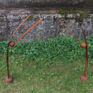 Pode incluir: Duas esculturas abstratas de metal com acabamento castanho enferrujado. Cada escultura tem uma base arredondada, um caule curvo, uma sec&ccedil;&atilde;o m&eacute;dia esf&eacute;rica e dois elementos delgados e curvos para cima. As esculturas est&atilde;o sobre uma superf&iacute;cie gramada.