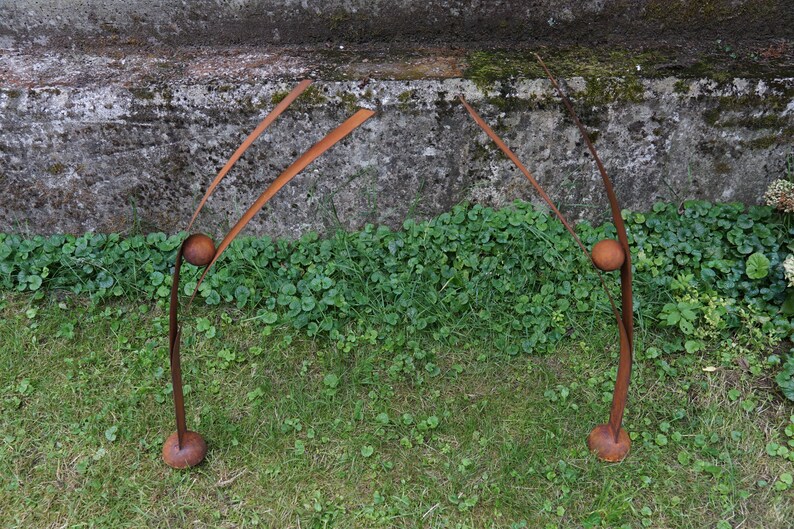 Pode incluir: Duas esculturas abstratas de metal com acabamento castanho enferrujado. Cada escultura tem uma base arredondada, um caule curvo, uma sec&ccedil;&atilde;o m&eacute;dia esf&eacute;rica e dois elementos delgados e curvos para cima. As esculturas est&atilde;o sobre uma superf&iacute;cie gramada.