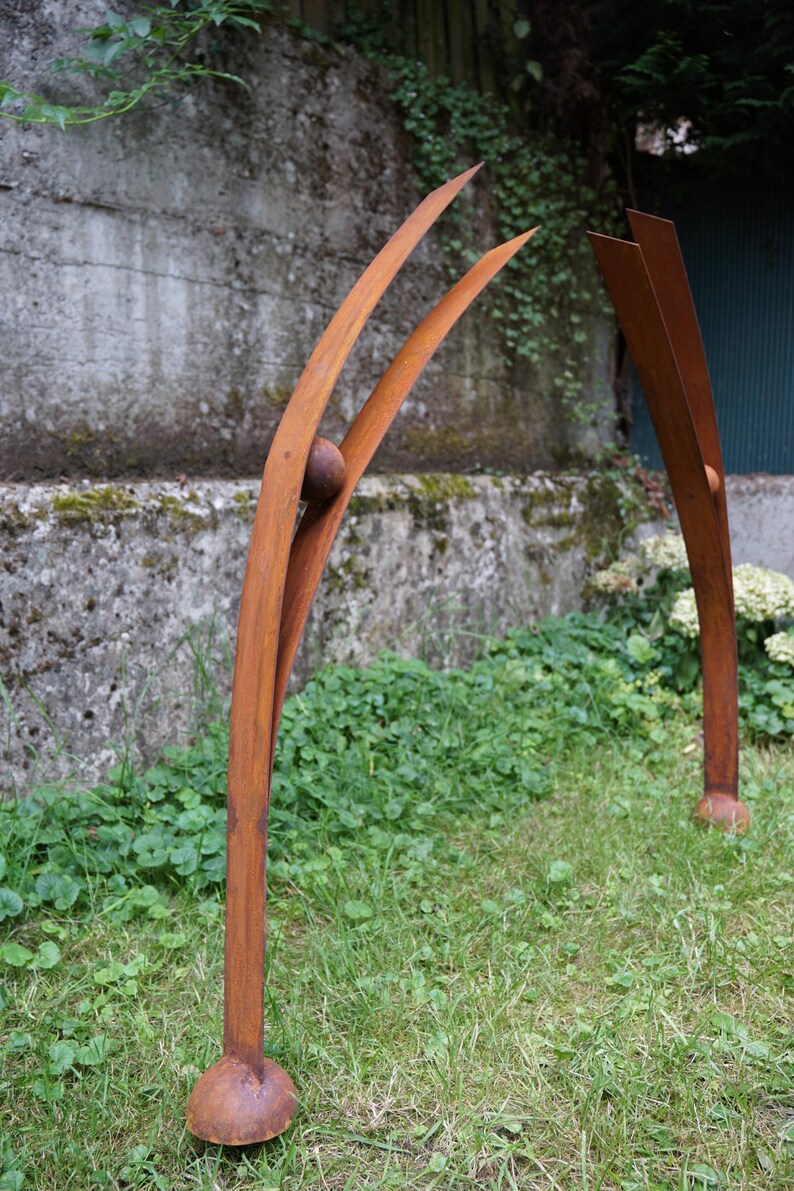 Pode incluir: Duas esculturas abstratas de metal com acabamento castanho-avermelhado desgastado. Uma apresenta dois elementos curvos e pontiagudos com uma esfera, a outra dois elementos retos e verticais. As esculturas est&atilde;o situadas numa &aacute;rea relvada, com um muro de bet&atilde;o ao fundo.