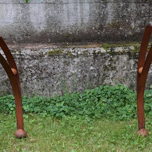 Pode incluir: Duas esculturas abstratas de metal enferrujado est&atilde;o em uma &aacute;rea gramada. Cada escultura tem um caule curvo com duas extens&otilde;es em forma de l&acirc;mina voltadas para cima e uma pequena esfera redonda. O fundo apresenta uma parede de concreto desgastada e folhagem verde.