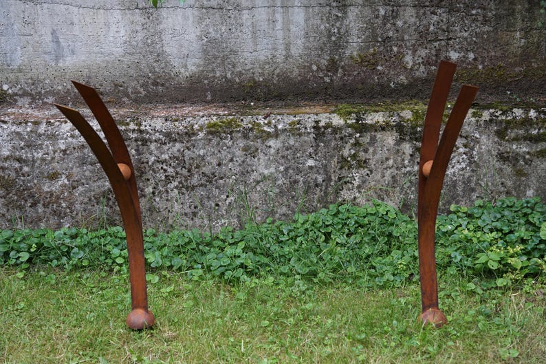 Pode incluir: Duas esculturas abstratas de metal enferrujado est&atilde;o em uma &aacute;rea gramada. Cada escultura tem um caule curvo com duas extens&otilde;es em forma de l&acirc;mina voltadas para cima e uma pequena esfera redonda. O fundo apresenta uma parede de concreto desgastada e folhagem verde.