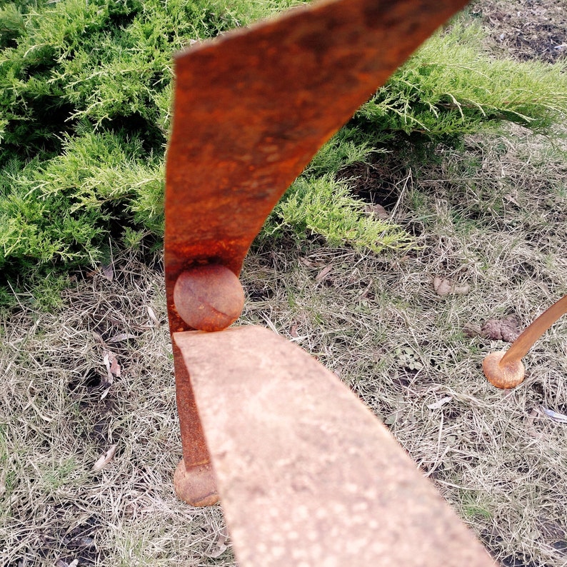 Pode incluir: Close-up de uma escultura de metal enferrujado e desgastado. A escultura apresenta elementos curvos em forma de l&acirc;mina conectados por detalhes esf&eacute;ricos. A pe&ccedil;a &eacute; colocada em um fundo de folhagem verde e grama seca, mostrando sua textura envelhecida.