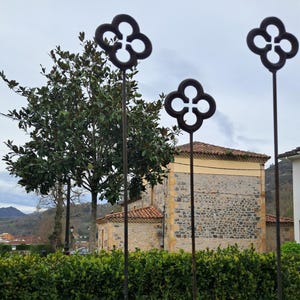 May include: Three wrought iron decorative stakes with a four-leaf clover design stand in front of a stone building with a red tile roof. A large tree and green bushes are in the foreground, with a cloudy sky in the background.