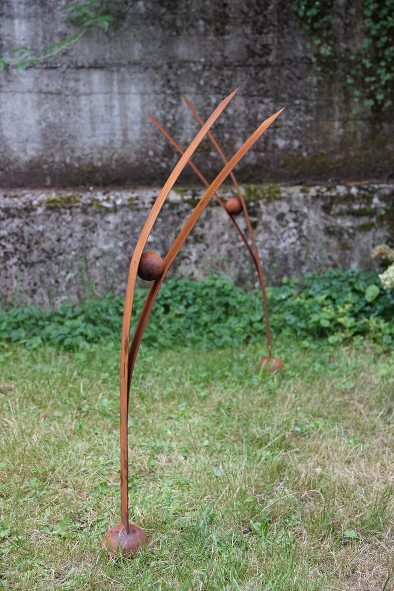 Pode incluir: Escultura abstrata em metal para exterior. Tr&ecirc;s finos caules arqueados de metal castanho com detalhes esf&eacute;ricos est&atilde;o na relva. A escultura est&aacute; em frente a uma parede de bet&atilde;o. O design &eacute; minimalista e abstrato.