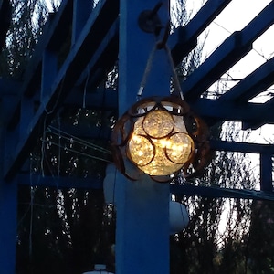 Könnte beinhalten: Eine hängende Außenleuchte mit einer kugelförmigen Glaskugel. Die Kugel ist in einem dekorativen Metallrahmen eingeschlossen und wird von einem Seil gehalten. Das Licht wird von warmen, gelben Lichtern beleuchtet, die eine gemütliche Atmosphäre schaffen. Die Leuchte hängt an einer blauen Holzkonstruktion.