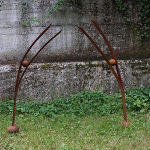 Pode incluir: Duas esculturas de metal para exterior com acabamento castanho enferrujado. Cada escultura tem um design curvo e arqueado com um acento esf&eacute;rico. As esculturas est&atilde;o colocadas num relvado gramado contra uma parede de bet&atilde;o texturizado.
