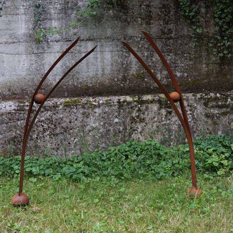 Pode incluir: Duas esculturas de metal para exterior com acabamento castanho enferrujado. Cada escultura tem um design curvo e arqueado com um acento esf&eacute;rico. As esculturas est&atilde;o colocadas num relvado gramado contra uma parede de bet&atilde;o texturizado.