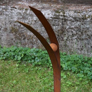 Pode incluir: Escultura de metal castanho enferrujado com uma esfera central e duas l&acirc;minas curvas para cima. A escultura est&aacute; sobre relva, com uma parede de bet&atilde;o ao fundo.