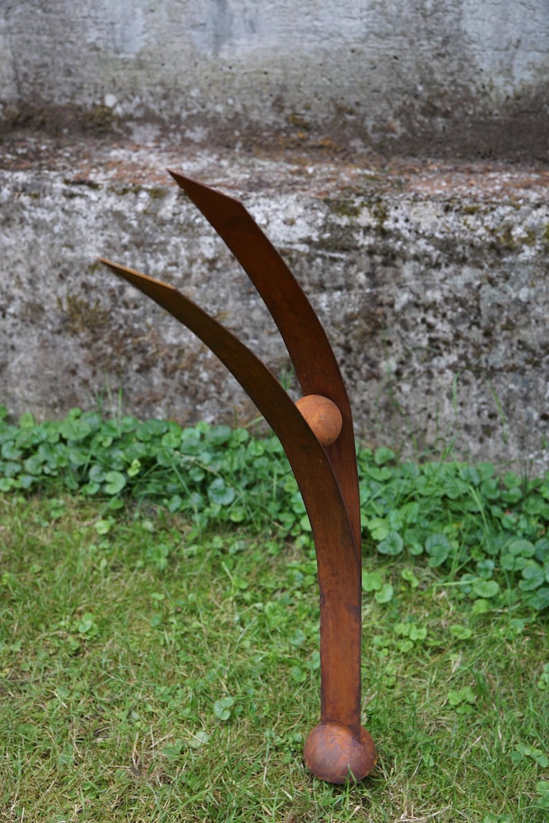Pode incluir: Escultura de metal castanho enferrujado com uma esfera central e duas l&acirc;minas curvas para cima. A escultura est&aacute; sobre relva, com uma parede de bet&atilde;o ao fundo.