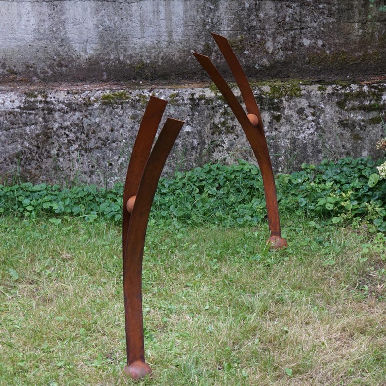 Pode incluir: Duas esculturas abstratas de metal com acabamento castanho enferrujado est&atilde;o sobre um relvado. Cada escultura tem elementos verticais curvos com detalhes arredondados e uma base. O fundo apresenta uma parede de bet&atilde;o e vegeta&ccedil;&atilde;o.