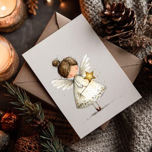 May include: A greeting card featuring a watercolor illustration of an angel holding a golden star. The angel is dressed in a white dress with delicate wings. The card is on a brown envelope, with candles and pinecones in the background.