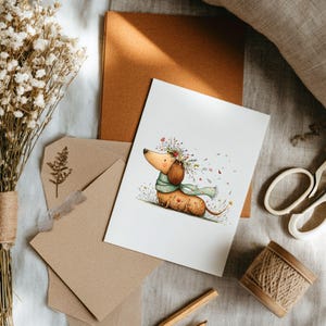 May include: A greetings card with a watercolour illustration of a dachshund wearing a flower crown and scarf. The card is on a table with stationery, dried flowers, and twine.