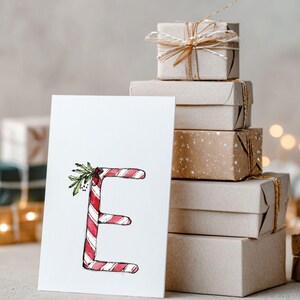 May include: A festive Christmas card with the letter 'E' styled as a candy cane, with red and white stripes and sprigs of holly. A stack of wrapped gifts in brown paper, tied with twine, is in the background, evoking a holiday atmosphere.
