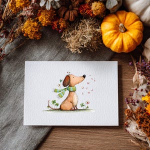 May include: A watercolour illustration of a dachshund wearing a green scarf, surrounded by pink petals and clovers. The dog is on a white card, with autumn foliage and pumpkins in the background. The artwork has a whimsical, autumnal theme.