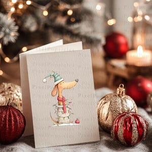 May include: A folded greeting card featuring a watercolor illustration of a tan dachshund wearing a teal striped hat and a pink scarf, sitting inside a white bucket wrapped in colorful Christmas lights, set against a textured paper background.
