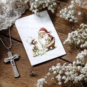 K&ouml;nnte beinhalten: Eine silberne Kreuzkette mit einem detaillierten Kruzifix liegt auf einer Holzoberfl&auml;che. Eine Weihnachtskarte mit Weihnachtsmann und Rentier ist ebenfalls vorhanden. Wei&szlig;e Blumen und Spitze erg&auml;nzen die festliche Szene.