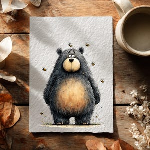 May include: A charming illustration of a bear looking concerned, with bees buzzing around it. The bear is depicted with black fur, a tan belly, and a light brown snout. The artwork is on textured paper, with a wooden background and a coffee mug.
