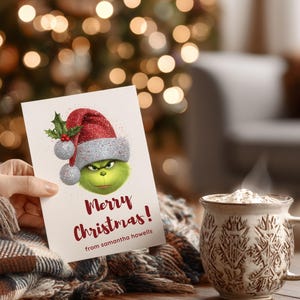 Peut inclure: Une carte de No&euml;l avec le Grinch portant un chapeau de P&egrave;re No&euml;l, avec le texte "Merry Christmas!" en rouge. Une tasse de boisson chaude est &agrave; c&ocirc;t&eacute; de la carte. La carte est tenue par une personne.
