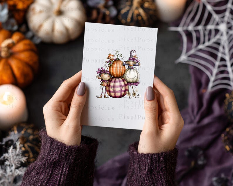 May include: A white rectangular card featuring a whimsical illustration of three stacked pumpkins with faces, including a central orange pumpkin, a bottom purple plaid pumpkin, and a white pumpkin on the right, all adorned with small decorative hats.