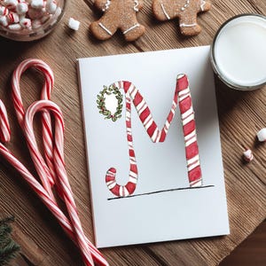 May include: A Christmas-themed image showing a white card with a candy cane-striped letter "M" decorated with a wreath. Candy canes, gingerbread biscuits, marshmallows, and a glass of milk are arranged on a wooden surface.