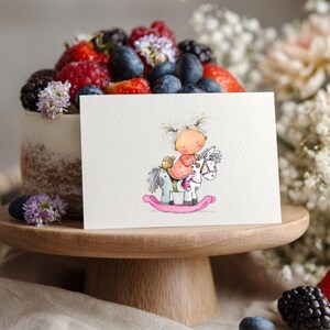 May include: A greeting card featuring a watercolor illustration of a child riding a rocking horse. The card is placed in front of a cake topped with berries and flowers, on a wooden cake stand. The scene is decorated with flowers and berries.