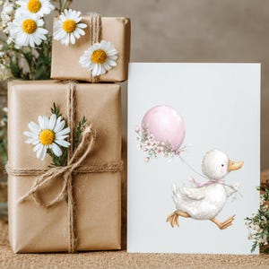 May include: A watercolour illustration of a white duckling running with a pink balloon. The image is on a white card next to wrapped brown paper gifts decorated with daisies. A vase of flowers is in the background.