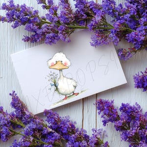 May include: A greeting card featuring a cartoon duck holding a bouquet of flowers, with a blue ribbon. The card is surrounded by purple flowers. The duck has a white body, a yellow beak, and pink cheeks. The background is a white wooden surface.