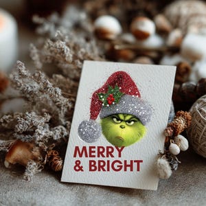 Peut inclure: Carte de v&oelig;ux de No&euml;l avec un personnage vert portant un chapeau rouge et argent&eacute; &agrave; paillettes. La carte porte l'inscription "MERRY & BRIGHT" en rouge. La carte est sur une surface neutre avec des d&eacute;corations festives.